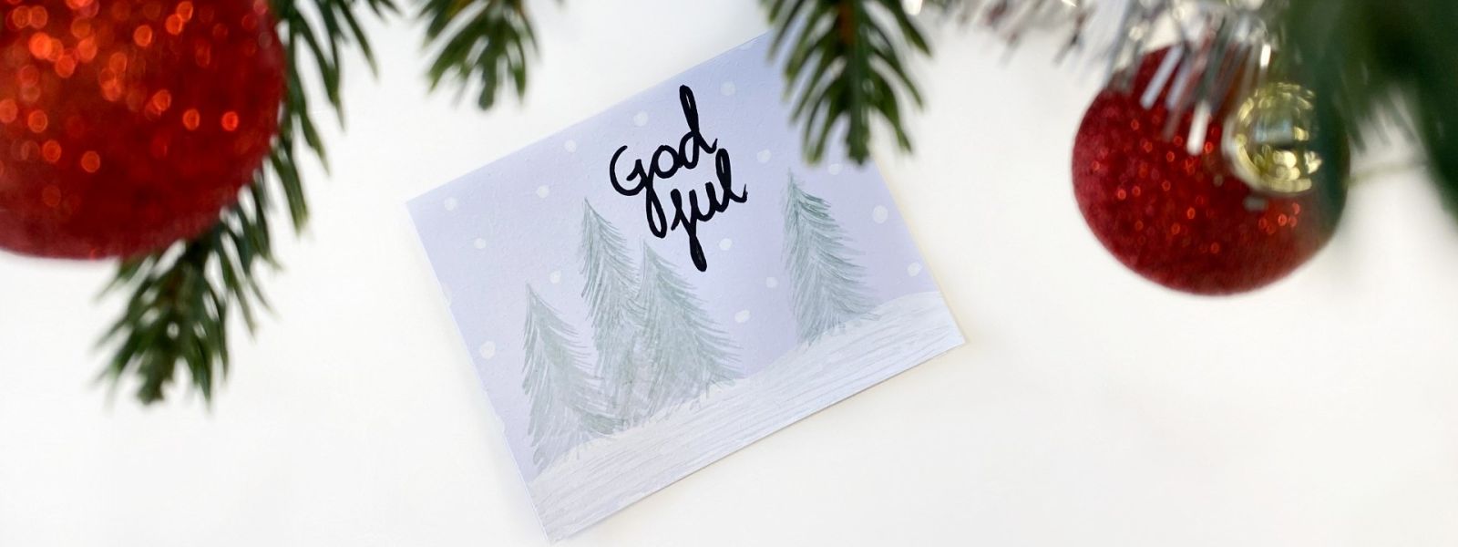 Närbild av julgran med röda glittriga kulor och ett kort med texten God jul framför snötäckta träd